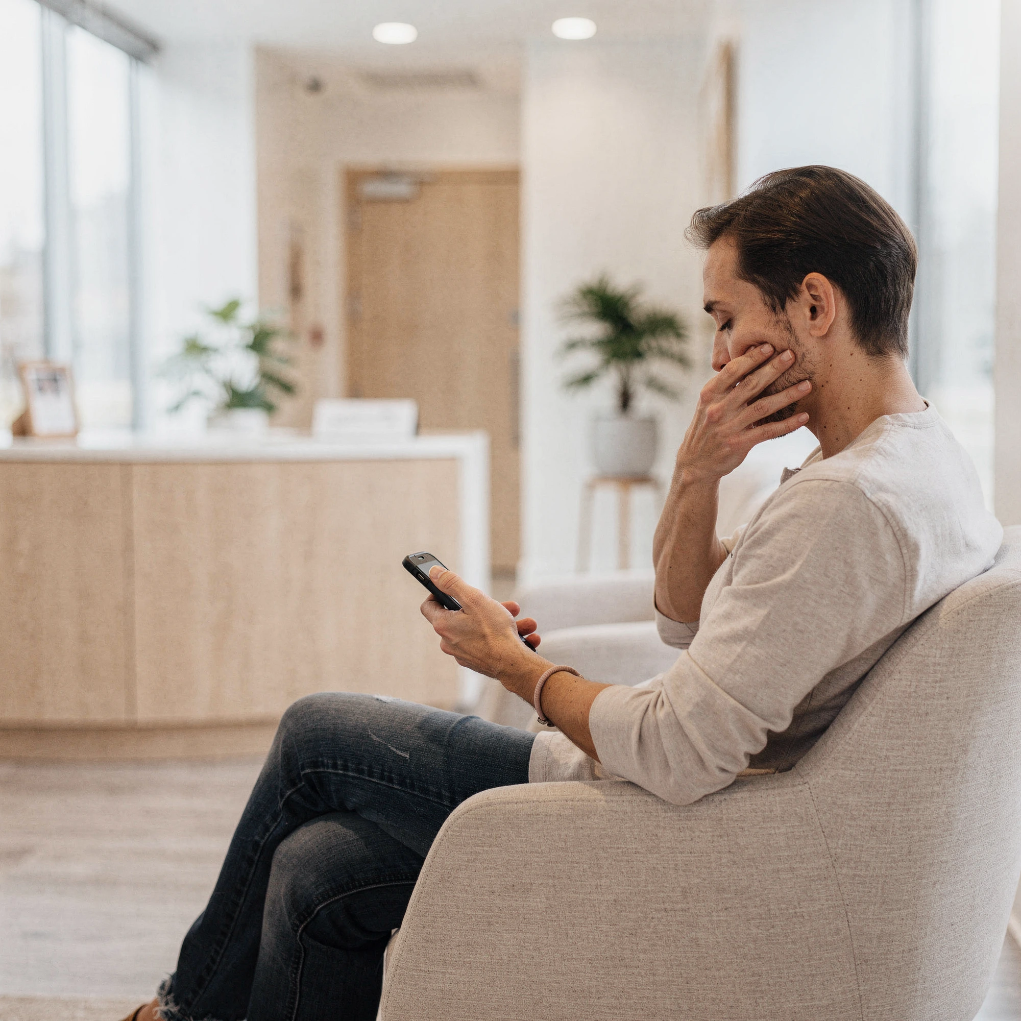 Man in dental waiting room holding phone and touching his cheek in discomfort